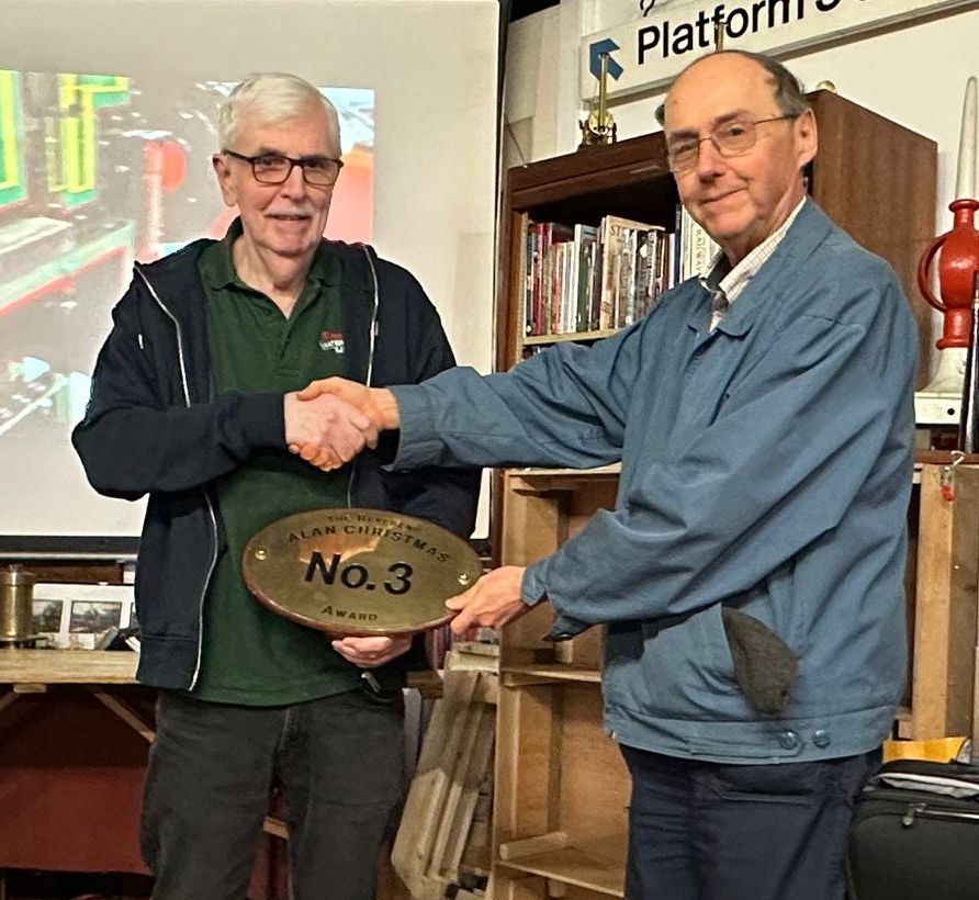 Frank Taylor presenting the Society's Reverend Alan Christmas Award to Richard Fillmore at the SWRS AGM in April 2024. Frank was himself a recipient of the award, in 2021. Standing in the SWRS Manor meeting place, surrounded by railwayana, Frank Taylor presents the replica brass oval cab numberplate that is the Society's Reverend Alan Christmas Award, to Richard Fillmore.