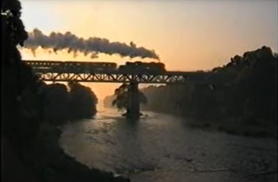 A steam-hauled charter run in June 2000 in the Czech Republic. – Ian Silvester – (still from YouTube video) A steam-hauled passenger train seen in silhouette crossing an under-slung truss girder bridge over a substantial river, at dusk.
