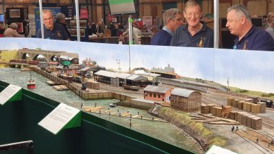A portion of the OO-scale layout 'Redbridge Wharf': a model of the sleeper works and associated timber wharf on the River Test, near Southampton; exhibited by Winchester Railway Modellers at the London Festival of Railway Modelling, Alexandra Palace, 19th March 2022 – Peter Jewell Exhibition-standard model railway featuring an extensive timber wharf and processing yard, with happy operators looking down from behind.