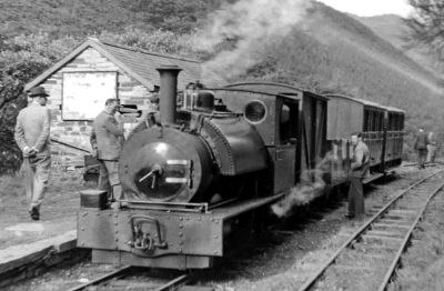 With mountainous scenery in the background, a small, narrow gauge steam locomotive waits by a low station platform with a short passenger train. An archive view of the early days of preservation on the Talyllyn Railway.