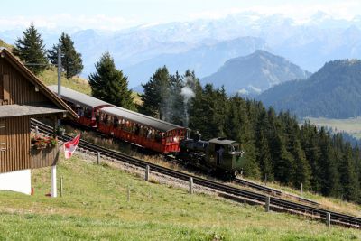 Preserved VRB H 2/3 standard gauge rack railway locomotive no. 16 (SLM No.2871, built 1923) with a train of 4-wheel coaches on the Vitznau-Rigi Bahn. The VRB climbs Mount Rigi by Lake Lucerne in Switzerland – 22 September 2007 – User:aforero – Wikimedia Commons CC-by-2.0 A small steam locomotive pushes two red, wooden, 4-wheel carriages up a steep rack railway on a grassy mountain slope. To one side is a traditional wooden Swiss chalet, complete with window box of flowers and Swiss flag. On the far side of the track is a grove of pine trees, with a deep, wide valley beyond, and a continuous backdrop of hazy mountain peaks.