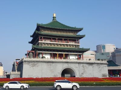 A traditional, and very old, Chinese temple with modern concrete buildings either side. A road runs past the front of the temple. The two are separated by a wide verge carpeted with colourful flowers.
