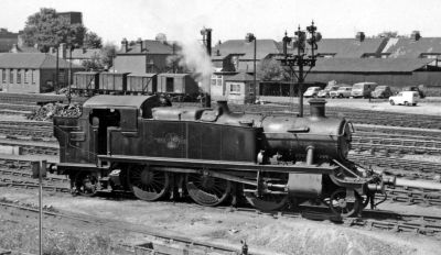 An archive view of Slough from June 1962. A lone 2-6-2T steam locomotive gently simmers with steam coming from its safety valve bonnet. Behind the locomotive are the many tracks of the loco shed, the Windsor Branch, and the Great Western Main line. In the background, a line of 12-ton vans rest in the goods sidings that run along the back of a row of 1930s housing.