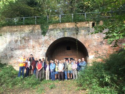 Railway Ramblers pose for a group photo in front of the long-closed Tyler Hill Tunnel on the former Canterbury to Whitstable line – October 2017. – c/o Keith Lawrie A group of 20-30 ramblers stand in front of the arched mouth of a disused single-track railway tunnel set in a tall, wide brick wall with tree foliage across the top and on either side.