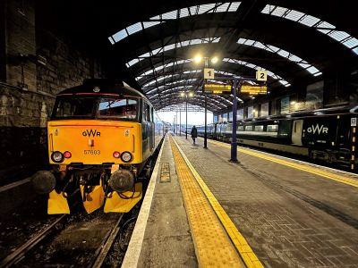A view along the platform inside a stone-built Victorian terminus station with a curved overall roof. A diesel locomotive sits at the head of the recently arrived sleeper service from London.