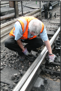 Frank Taylor demonstrating use of a point clip to secure a set of points (or 'switch'), to prevent them from being changed remotely. A railway worker, wearing an orange high-visibility vest, crouching down to clamp the moving rail of a point (the switch rail) to the fixed (or 'stock') rail using a point clip