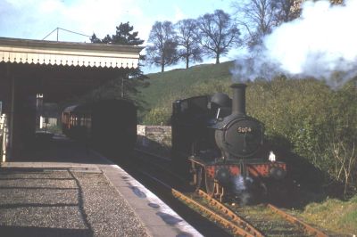 A small steam locomotive (Class 58xx 0-4-2T) runs round its train at Tetbury station in 1962. The decorative valance of the platform canopy casts a deep shadow over the single passenger coach. Trees line the top of the grassy embankment behind, emphasising the rural location.