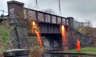 The 125 year-old bridge carrying the Great Central Railway over the Grand Union Canal at Loughborough. On 12th December 2018, contractors are cutting free the lattice bridge parapets so they can be restored off-site. (courtesy Tom Ingall) A traditional plate steel girder bridge, with engineering brick abutments, built to carry a railway across a canal. Two showers of sparks are seen falling from the side of the bridge as contractors cut away the steel lattice work parapets.