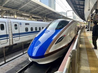 E7 series Shinkansen train at Tokyo (Central) Station ready for a service on the Tokyo-Nagano route – April 2023 (courtesy Chris Zaremba) A close-up of the cab end of a highly-streamlined express passenger train in a city station in Japan. The high-gloss finish of the bodywork is very obvious.