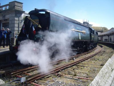 Un-rebuilt Bulleid Pacific 34067 'Tangmere' at Folkestone Harbour, in a recreation of the 'Golden Arrow' train. – (courtesy Dick Crane) A streamlined 4-6-2 steam locomotive in the sharply curved platforms of the closed Folkestone Harbour station. The 'Battle of Britain' class locomotive is partly obscured by steam, and features large, three-dimensional gold arrows on the front and side.