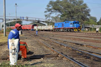 A Zambian rail worker sits patiently by a point lever, waiting for his next task. Beyond an extensive track layout, a modern diesel locomotive sets off with a mixed train of freight cars and passenger coaches.