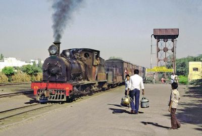 A somewhat run-down narrow gauge tender steam locomotive draws a train of elderly wooden carriages into a wayside station in rural India. A railway worker carries cans of water across the ground level platform, towards the train.