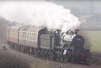 With a plume of exhaust steam trailing up and behind, a small GWR tender engine hauls a passenger train on the West Somerset Railway in 2022.