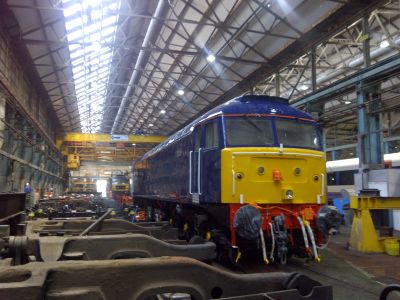 Unidentified Class 47 under overhaul at Eastleigh Works on 20th October 2018 – Peter Greatorex Interior view of an early 20th century locomotive workshop, complete with overhead crane. A freshly painted Class 47 diesel locomotive sits alongside components of other rolling stock.
