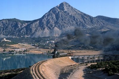 Rural Turkey: the lower slopes of a mountain range are covered by a scattering of houses surrounded by many trees. Against this backdrop, a large steam locomotive is hauling a mixed train of one passenger coach and two vans onto an embankment alongside a substantial inland lake, having just crossed a spindly metal viaduct. The arid appearance of the embankment and fields around the lake is a stark contrast to the abundant woodland beyond.