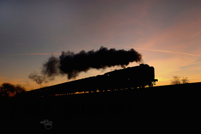Photo copyright: Jack Boskett A large steam locomotive hauling a passenger train along an embankment, silhouetted against a sunset sky
