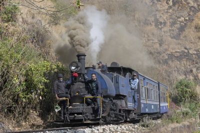 Darjeeling Himalayan Railway B-Class locomotive 01B on a downhill service near Mahanadi, 16th January 2024. – Simon Colbeck A long but low saddle tank locomotive pulling a passenger train on the Darjeeling Himalayan Railway. The bright blue livery of the dirty locomotive and its two short coaches contrasts starkly with the scrubby surrounding vegetation clinging to the sheer rock face beside the line. Two railway workers are standing on the front of the engine, watching the track ahead, while the driver and fireman are looking out from the cab.