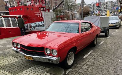 1970 Chevrolet Chevelle... plus trailer! – Rob Holder A bright red American sports coupe from 1970, parked on a harbour-side road amid brick-faced apartment blocks in a European city.