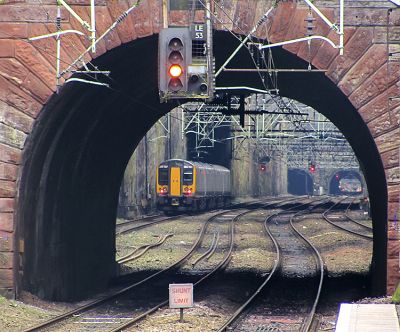 A neatly dressed stone arch bridge frames the view along the track into a deep cutting beyond. A four car electric multiple unit is dwarfed by its surroundings. Four tracks snake away towards two arched tunnel mouths in the distance. Above the tracks is a jumble of bracketry for the overhead wires.
