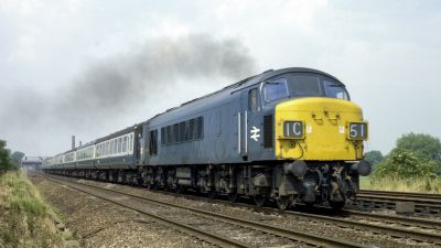 A very long British Rail diesel locomotive, painted BR Blue, with yellow ends, hauls an express passenger train through flat countryside.