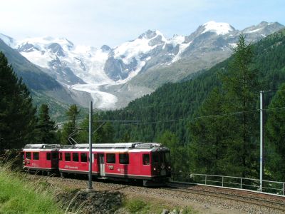 A boxy, but colourful, electric multiple unit on a steep climb in Switzerland, backed by dense forest and a glacier in the background.
