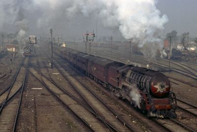 A mainline railway scene in India in the 1980s. A large, partly-streamlined steam locomotive hauls a lengthy passenger train through broad expanses of freight marshalling yard, between characteristic semaphore signals. Smoke from several locomotives contribute to the heavily polluted sky.