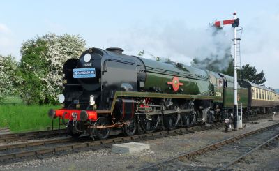 A large steam locomotive with smoke deflectors, in shiny ex-works condition, hauls a passenger train past a semaphore signal on a rural heritage railway.