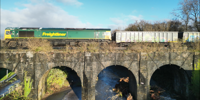 A modern diesel locomotive hauls a train of bogie mineral wagons over an almost 200-year-old stone, arched river bridge.
