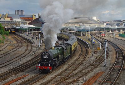 A large, preserved steam locomotive hauling an excursion train threads its way along the twisty tracks leading away from a major city station.