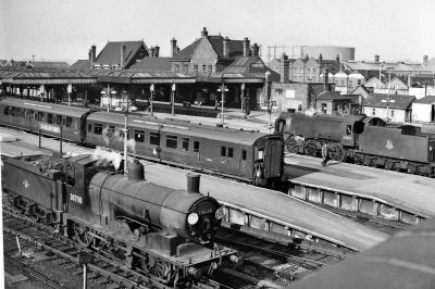 Guildford station in the 1950s, showing the Victorian station building and many platforms and canopies, with the town, and gas works, beyond. An electric multiple unit sits at one platform, and a couple of 0-6-0 tender steam locomotives, spanning 50 years of loco design, rest on nearby tracks.