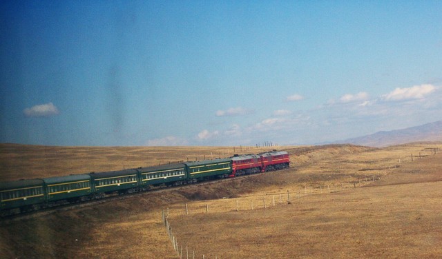 A double-headed passenger train crossing a barren landscape under clear blue skies.