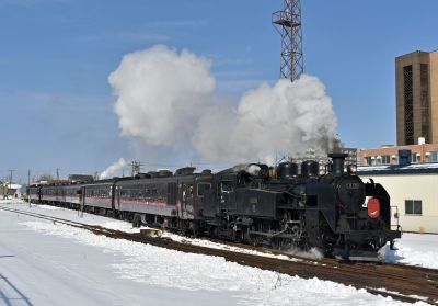 A vintage Japanese steam locomotive hauls a lengthy passenger train through an urban station in snowy conditions.