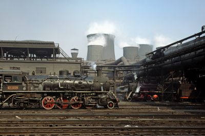 Two medium-large Chinese steam locomotives in an industrial setting. An array of cooling towers in the background identify the location as a power station.