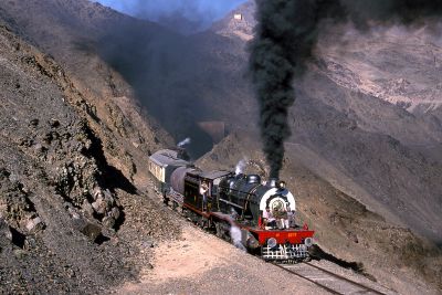 A short passenger train, with large steam locomotives working hard at front and rear, emerges from a tunnel into a barren, mountainous landscape in Pakistan, without a trace of vegetation anywhere.