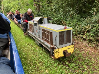 A diesel locomotive hauls excited passengers on a miniature railway train through woodland.