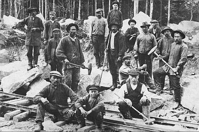 A group of navvies, in tough work clothes, flat caps and boots, tools in hand, stand and sit around a length of railway under construction. (Finland, 1918).