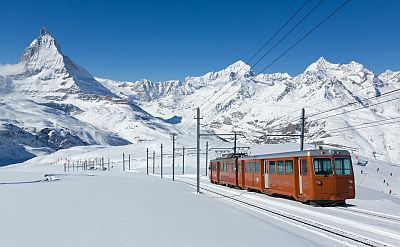 Under a deep blue sky, high in the Swiss mountains, the dark orange livery of a 2-car electric train stands out against the bright white background of untouched snow fields and the mountain peaks behind.
