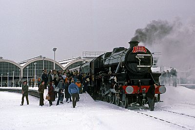 In a landscape of freshly fallen snow, a newly restored steam locomotive stands at the head of an excursion train at a major city station. A throng of eager enthusiasts fills the platform by the engine.