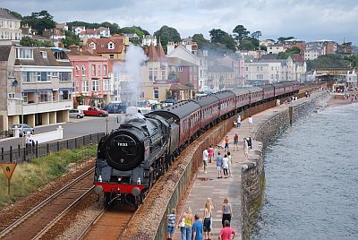 A large steam locomotive hauls a train of contemporary coaching stock through Dawlish. To the left and behind are row upon row of assorted Victorian guest houses and hotels. To the right is the stone-built sea wall, and then, as its high tide, the sea!