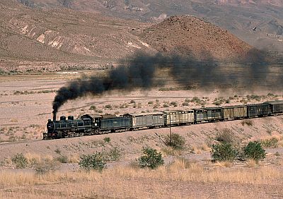 A steam-hauled mixed (passenger and freight) train charges across a plain, somewhere in South America, dwarfed by arid mountains in the background.