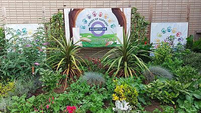 A small, densely-packed flower garden; the Elizabeth line roundel on the wall behind revealing that this is at West Ealing station.