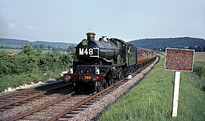 An express passenger steam locomotive, polished up for the occasion and with a suitable headboard, hauls the last steam-hauled passenger service from Paddington on a double-track mainline through a wide expanse of British countryside, with low hills in the background. Many enthusiasts are hanging out of the windows to savour the experience.