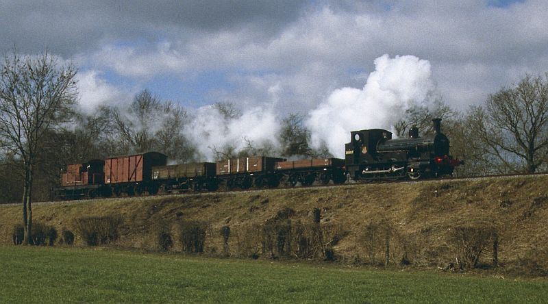 A small 2-4-0WT steam locomotive (a Beattie Well Tank) hauls a short freight train across an embankment on a rural heritage railway in the south of England.