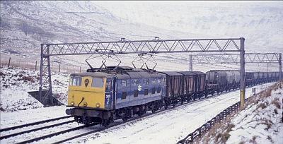 Snowy view of an early British Railways design electric locomotive hauling a freight train formed of short wheelbase vans, grain wagons, and empty coal hoppers, 'under the wires' on the Woodhead line.