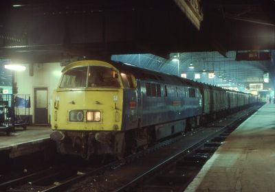 A night time scene at a major London railway terminus station in the 1970s. A diesel-hydraulic locomotive sits at the front of a rake of parcels vans.