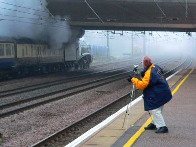 In the background, a mainline steam locomotive (A4 Pacific 60019 Bittern) pulls a passenger train away from a major station. On the empty platform opposite is a lone trainspotter, complete with long anorak, hoping to capture the scene on his tripod-mounted camera.